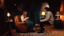 a man sits on a stool next to a woman who says you 're such an interesting young guy in a dark room