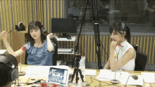 two women wearing headphones are sitting at a table with a laptop and a microphone