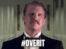 a man in a tuxedo with a mustache is standing in front of a wall and says #overit .