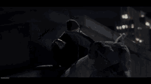 a man in a black hat is standing on a rooftop at night looking at his watch