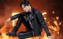 a man in a leather jacket sits on the ground in front of a fire background