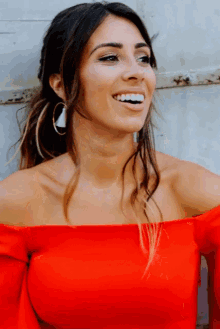 a woman wearing a red off the shoulder top and hoop earrings smiles