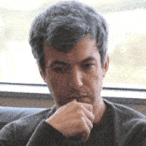 a man with gray hair is sitting in front of a window with his hand to his chin