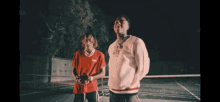 two young men are standing next to each other on a tennis court at night .