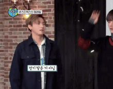 a man in a black jacket is standing in front of a brick wall while another man waves his hand in front of him .