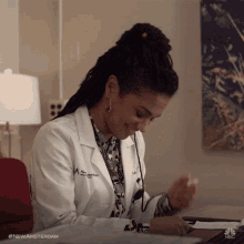 a woman in a lab coat is sitting at a desk with a nbc logo in the background