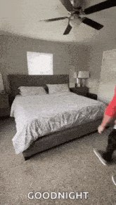 a man is standing next to a bed in a bedroom with a ceiling fan and a window .
