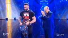 a man in a we the people shirt is holding a wrestling championship belt while standing next to another man .