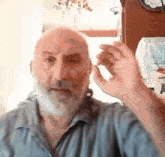 a bald man with a beard holds his hand to his ear while wearing a blue shirt