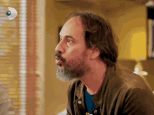 a man with a beard is sitting in front of a window with blinds and a blue circle with the letter d on it