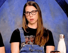 a woman wearing overalls and glasses is sitting in front of a blue wall