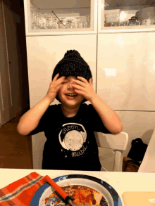 a young boy covering his eyes with his hands while wearing a shirt that says moon music