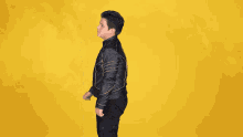 a man in a black leather jacket stands with his arms crossed against a yellow background