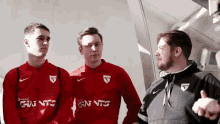 a man wearing a black nike jacket talks to two guys wearing red shirts that say gn ntc