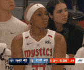 a female basketball player wearing a mystics jersey sits in the stands