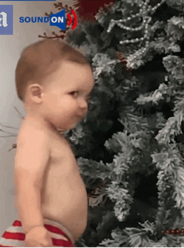 a baby is standing in front of a christmas tree with a soundon logo in the corner