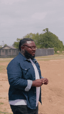 a man in a denim shirt and glasses is standing in a dirt field