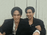 two men are sitting next to each other in front of a white wall and smiling .