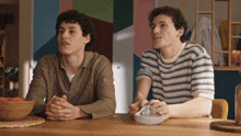 two men are sitting at a table with a bowl of oranges on it