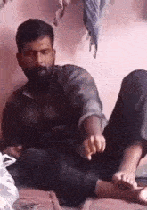 a man with a beard is sitting on a bed with his feet up and pointing at something .