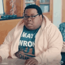 a fat man wearing glasses and a pink hoodie is sitting at a table with his hands on the table .