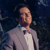 a man wearing a purple suit and a pink bow tie smiles with his eyes closed