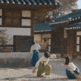 three women are sweeping the ground in front of a building with a blue roof