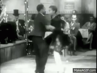 grayscale GIF of a well-dressed, black man twirling a well-dressed, black woman while they dance in a 1920s speakeasy