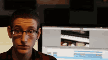 a man wearing glasses looks at the camera in front of a computer monitor