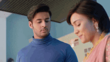 a man in a blue turtleneck sweater stands next to a woman in a pink dress