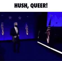 a man in a suit and tie is walking on a stage with the words hush , queer ! above him .