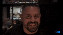 a man with a beard is smiling in a dark room with a blue infinity symbol in the corner