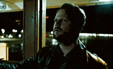 a man with a beard and a leather jacket is standing in front of a window at night .