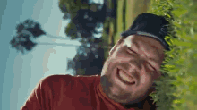 a man wearing a red shirt and a hat is laughing while leaning against a tree .