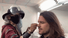 a man with long hair is getting his face painted by a woman wearing a face mask