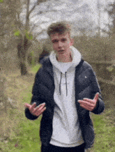 a young man wearing a black jacket and a white sweatshirt is standing in a field with his hands outstretched .