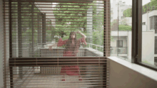 a woman in a red dress is standing in front of a window with blinds on it