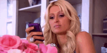 a woman is holding a cell phone in her hand while sitting in front of a bouquet of pink roses .