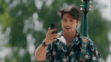 a man in a flamingo shirt is looking at his cell phone while standing next to a guitar .