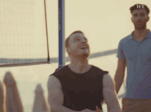 a man in a black tank top is laughing while a man in a blue shirt stands nearby