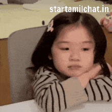 a little girl in a striped shirt is sitting at a table with her head resting on her arms .