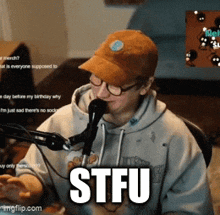 a man wearing a hat and glasses is sitting in front of a microphone with the text stfu above him