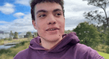 a young man in a purple hoodie looks at the camera with trees in the background