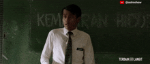 a man in a white shirt and tie stands in front of a blackboard that says " kemahiran hidup "