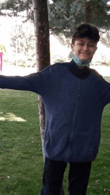 a young boy wearing a mask and a blue sweater stands in front of a tree