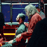 a man with a beard sits in a dark room with a red jacket on