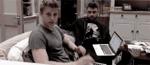 two men are sitting on a couch with laptops and one of them is wearing a shirt that says ' ?? ' on it