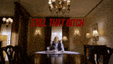 a man sits at a table in a room with the words still that bitch above him