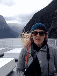 a woman wearing sunglasses and a knitted hat stands in front of a body of water
