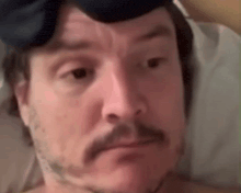 a man with a mustache is laying in bed with a headband on his head and making a funny face .